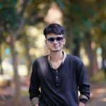 Stylish young man wearing sunglasses and black attire, standing confidently on a forest path.